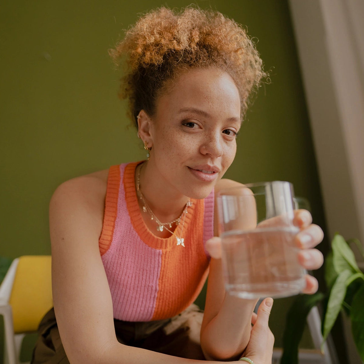Een vrouw in een roze topje die een glas water vasthoudt, in een huiselijke omgeving met planten.